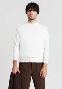 Weißer Rundhalsausschnitt-Pullover aus weichem Stoff, mit langen Ärmeln und gerippten Bündchen. Getragen mit dunkelbraunen, gefalteten Hosen.
