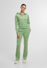 Világos zöld bársonynadrág, zippzáras kapucnival, elülső zsebekkel és szűk szabású nadrággal. Kiegészítők között arany színű fémalkatrészek és zsinórok találhatók.