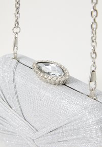 Borsa a mano in tessuto argento con un design plissettato, adornata con una gemma trasparente a forma di goccia e maniglie in argento, dotata di una tracolla a catena.