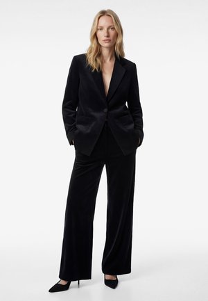 Femme blonde portant un costume en velours noir avec un pantalon à jambes larges et une veste, debout les mains dans les poches devant un fond uni.