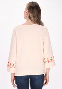 Hellbeige Oberteil mit weiten Ärmeln, floralen Akzenten an den Bündchen, glatter Textur und lockerer Passform. Abgerundet mit einem Rundhalsausschnitt.