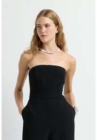 Mono negro sin tirantes con un cuerpo ajustado y piernas anchas, hecho de una tela suave; complementado con un collar de tono plateado.