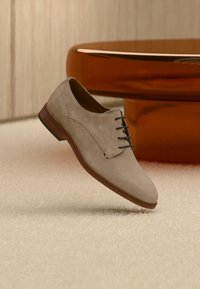 Μπεζ σουέντ παπούτσι με κορδόνια, μυτερή μύτη και καφέ δερμάτινη σόλα· διαθέτει διακοσμητικό ράψιμο και λεπτή λεπτομέρεια λογότυπου.