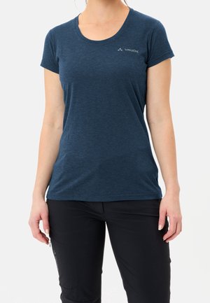 Kobieta ubrana w ciemnoniebieski krótki rękaw Vaude t-shirt i czarne spodnie, stoi z ramionami swobodnie opuszczonymi wzdłuż ciała na białym tle.