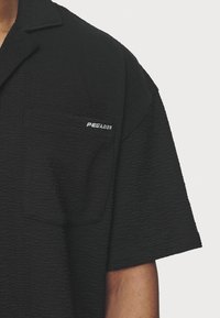 Camicia nera a maniche corte con trama testurizzata e un motivo sottile, con taschino sul petto ricamato con la scritta "PEGADOR" in bianco.