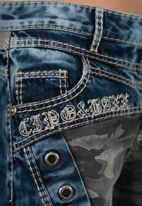 Jean en denim avec un lavage bleu, des coutures beige contrastantes et une poche en camouflage. Présente un logo de marque et des accents en métal.