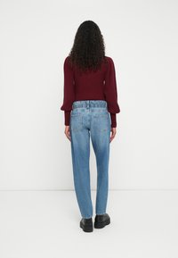 Ženska s kodrastimi lasmi stoji obrnjena stran, oblečena v temno rdečo dolgosrajčno pulover, svetlobluje visoko pasne jeans hlače in črne platform čevlje.