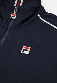 Marine blauwe zip-up jas van zachte stof, met witte en rode accentstrips en een logopatch met rode, witte en marine details.
