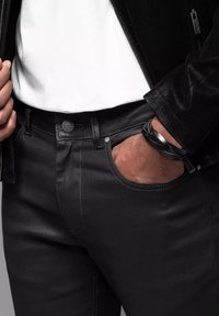 Vaqueros de mezclilla negros con un ajuste slim, con acentos de botones y remaches plateados, y costuras en gris en contraste. Llevado con una camisa blanca y una pulsera negra.
