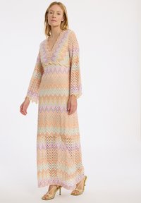 Robe maxi avec un motif en zigzag dans des couleurs pastel, dotée d'un décolleté en V profond et de longues manches évasées. Le matériau semble léger et texturé.