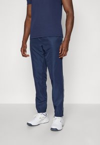 Pantaloni della tuta blu navy con strisce bianche ai lati, polsini elasticizzati e un tessuto leggero e morbido. Presentano tasche con zip e un dettaglio con logo.