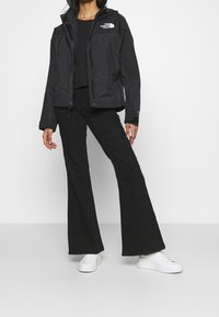 Svart vattentät jacka med full dragkedja framtill, justerbar huva och logotypaccent på vänster bröst, tillsammans med svarta utställda byxor och vita sneakers.