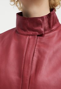 Κοντινό πλάνο ενός ατόμου που φοράει μπουφάν από μπορντό δέρμα με ψηλό γιακά, εμφανείς ραφές και λεία υφή.