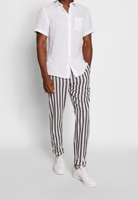 Man in een wit kortemouwen overhemd, zwarte en witte verticale gestreepte broek en witte sneakers, staande tegen een effen achtergrond.