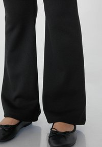 Schwarze, ausgestellte Hosen mit glatter Textur, kombiniert mit schwarzen Ballerinas, die vorne eine Schleife aufweisen. Nahtdetails am Saum.