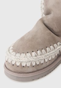 Stivale in suede grigio con punta tondeggiante, caratterizzato da cuciture intrecciate di colore crema lungo la parte superiore. Superficie testurizzata e suola in gomma.