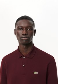 Burgundy pikétröja i texturerad tyg, med en tvåknappslång placket och en grön broderad logotyp på vänstra bröstet.