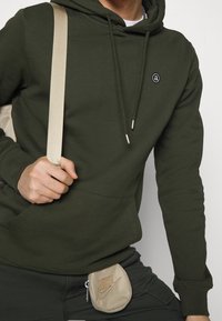 Felpa verde oliva con cordoncini e tasca frontale, abbinata a una pochette beige chiaro con logo Nike. Tessuto testurizzato, design casual.