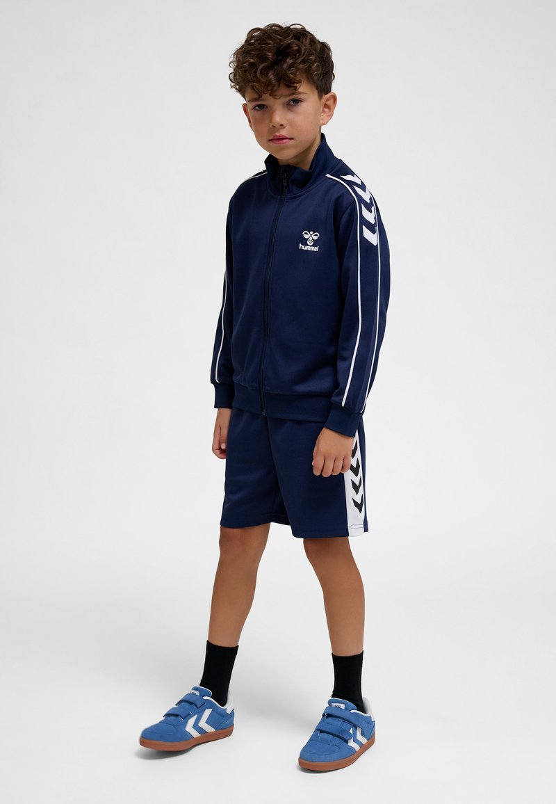 Fiú áll, tengerészkék sportdzsekit és rövidnadrágot visel, fehér csíkokkal, valamint kék sportcipőt fekete zoknival, egyszínű háttér előtt.