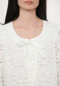 Bluză albă din dantelă, cu guler rotund și detalii cu fundiță. Prezintă modele florale, textură transparentă și închidere cu butoane în față.