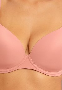 Soutien-gorge rembourré rose clair avec un tissu lisse, un design plongeant et des bretelles ajustables. Présente une finition sans couture et des détails de couture subtils.