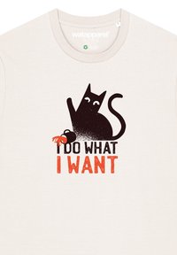Beige katoenen t-shirt met een grafiek van een zwarte kat met witte ogen. De tekst eronder luidt "IK DOE WAT IK WIL" in vetgedrukte oranje en zwarte letters.
