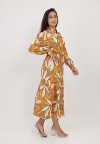 Abito floreale marrone a lunghezza media con maniche lunghe, motivi di foglie beige e arancioni, apertura con bottoni, vita cinched e gonna a strati. Sandali con tacco trasparente.