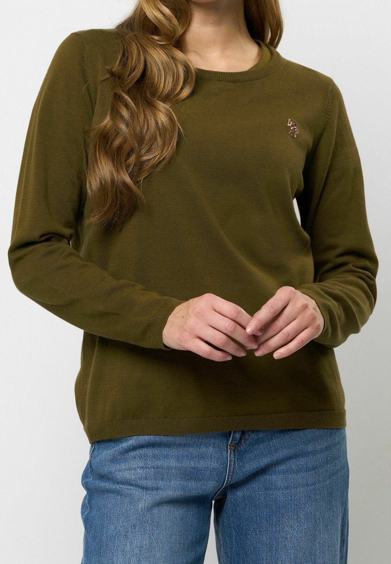 Femme portant un pull à manches longues vert olive avec un petit logo brodé sur la poitrine, associé à un jean bleu, mains doucement jointes.