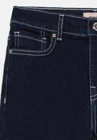 Mørkeblå denimbukser med kontrasterende hvit søm, frontlommer og sølvdetaljer. Har et klassisk fem-lomme-design.