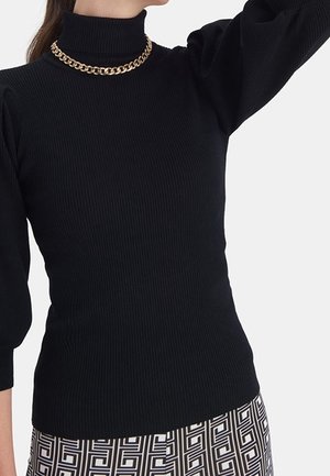 Kobieta w czarnym prążkowanym swetrze z golfem, złotym naszyjnikiem łańcuszkowym oraz spódnicy w geometryczny wzór czarno-białych kwadratów.