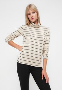 Beige csíkos garbó pulóver fekete csíkokkal; puha anyag, testhezálló fazon, háromnegyedes ujjak, ribbed fekete leggings-szel párosítva.