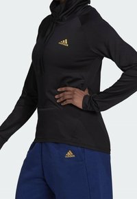 Personne portant un sweat à capuche noir à manches longues et un pantalon de survêtement bleu, tous deux avec des logos Adidas dorés, une main reposant sur la hanche.