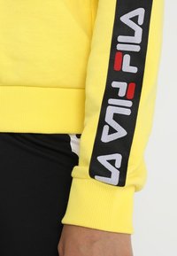 Sudadera amarilla con puños acanalados que presenta una raya lateral negra con bordado del logo "FILA" en blanco y rojo. Material de algodón suave y cepillado.