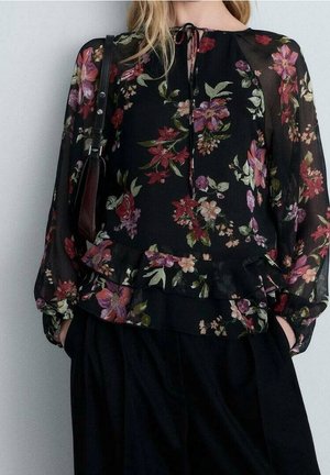 Vrouw draagt een zwarte doorschijnende bloemenblouse met lange mouwen, gerimpelde taille en een gebonden hals, gecombineerd met losse zwarte broek en een schoudertas.
