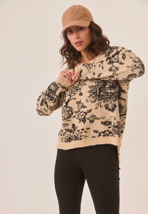 Suéter de punto beige con patrón floral negro, cuello redondo y puños acanalados, combinado con jeans negros y una gorra color beige.