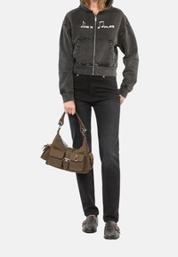 Grijze zip-up hoodie met grafische muzieknoot, zwarte jeans en donkere loafers. Draagt een bruine stoffen handtas met meerdere zakken en gespen.