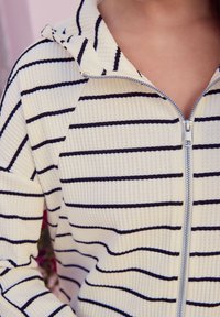 Gestreepte hoodie met ritssluiting in crème met marineblauwe horizontale strepen. Wafelgebreide textuur, capuchonontwerp en ritssluiting met metalen trekker.