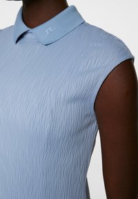Vaaleansininen hihaton polo-paita, jossa on tekstuurinen aaltoileva kuosi, pehmeä kaulus ja hillitty logo kauluksessa.