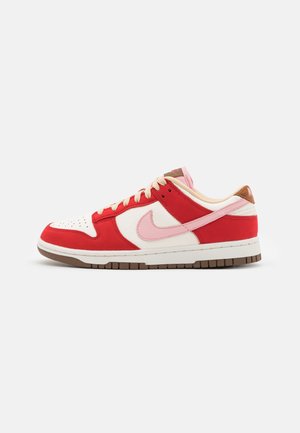 Baskets basses en cuir blanc et rouge avec logo swoosh rose, lacets beiges et semelle marron, présentées de profil sur un fond uni.