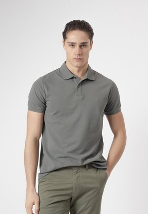 Joven con el pelo peinado hacia atrás, vestido con una camisa polo gris y pantalones verde oliva, de pie con una mano en el bolsillo sobre un fondo blanco.