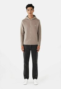 Lys beige zip-up hettegenser med fremre sømdetalj, som komplementerer svarte jeans og hvite joggesko i et avslappet antrekk.