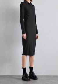 Schwarzes geripptes Kleid mit Kragen, langen Ärmeln und mittellangem Schnitt, kombiniert mit schwarzen Plateauschuhen und Socken. Strukturiertem Stoff.