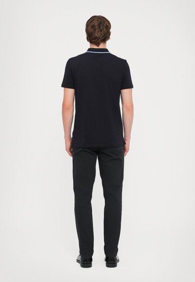 Camisa polo preta de mangas curtas com detalhe em colarinho azul claro, combinada com calças pretas de perna reta e sapatos pretos. Vista pela parte de trás.