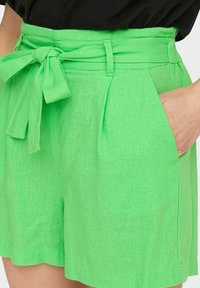 Pantalones cortos de lino verdes con una cintura alta, cinturón para anudar, pliegues y bolsillos laterales. La tela tiene un aspecto texturizado.