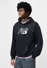 Μαύρο φούτερ με κουκούλα με μεγάλο, στυλιζαρισμένο ασημί λογότυπο New Balance. Διαθέτει μπροστινή τσέπη και στρωμένο μωβ πουκάμισο από κάτω. Μαλακή υφή.