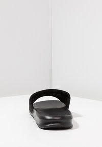 Tongs noires avec une finition mate lisse, large sangle ouverte et semelle légèrement surélevée. Semelle intérieure texturée pour un meilleur grip et un confort accru.