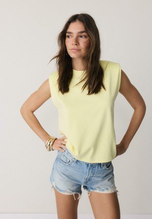 Mladá žena s hnedými vlasmi po ramená, oblečená v bezrukávovom svetložltom tričku, riflových šortkách a zlatých náramkoch, stojaca s rukami v bok.