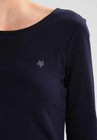 Navy blauwe langarmtop van gladde stof, met een subtiele grijze logo op de borst en een wijde halslijn.
