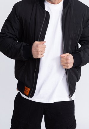 Homme portant une veste bomber noire par-dessus un t-shirt blanc, tenant la fermeture éclair de la veste avec les deux mains, sur un fond clair uni.