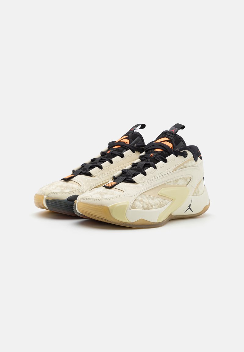 Zapatillas deportivas de color crema con paneles de malla, cordones negros y suelas de goma. Presentan un diseño anguloso y un logo en el lado exterior.
